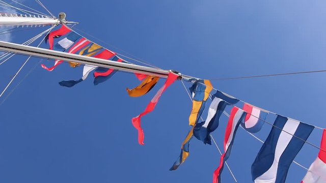 Upward shot of variety of colorful flags blowing in strong breeze on tall wooden ship with cloud and blue sky in background