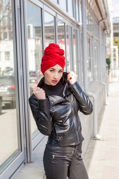 Stylish cheeky Muslim girl with a fashionably tied headscarf turban on her head in a red coat. Screaming image of a modern woman in the city against the backdrop of architecture