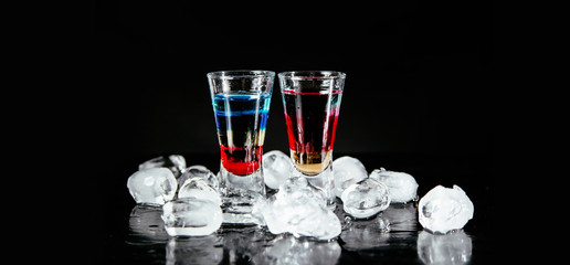 Two colorful alcohol shots on a black counter with ice next to them.