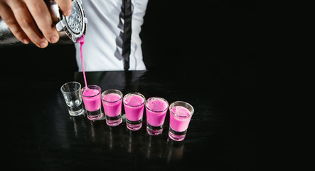 Barman making pink alcohol shots on a black counter. Bartender pours pink cocktail from a shaker into shots. © JustLife