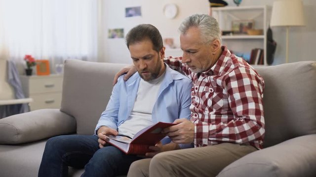 Pensive adult males holding album and viewing photos, recalling relatives