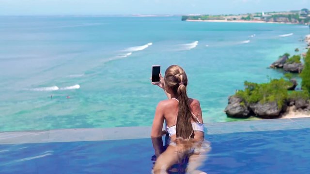 A young and beautiful girl in an open-air pool on a tropical island takes a smartphone camera landscape. Woman swimming in the pool overlooking the sea