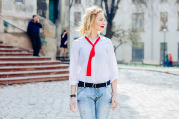 Fototapeta premium Beautiful blonde young woman red lips,fresh face wearing white shirt, red tie, blue jeans model, sensual woman walking on the street sunny spring day.