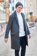 Young attractive hipster man wearing a gray coat, white sweater and black jeans walking in the street.