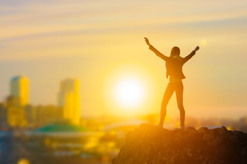Slender girl businessman on top of a mountain holding her hand up, against the background of the city in the rays of the sun. Business concept idea, happiness, success and achievement, leadership.
