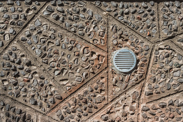 Silver round metal ventilation grille on a gray and orange concrete wall with stones background