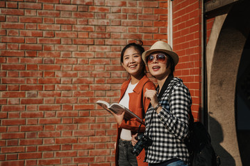 Fototapeta na wymiar two fashion asian women travelers in sunglasses and camera walking out finish visiting chinese temple by red brick wall copy space. young girls with guide book enjoy sunset sunshine smiling cheerful.
