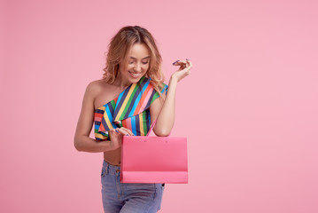 Fototapeta premium Portrait of an excited beautiful smiling girl wearing colorful clothes holding shopping bag isolated over pink background. Place to sign or text on the package