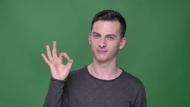 Closeup shoot of young handsome caucasian malegesturing ok handsign looking straight at camera with background isolated on green