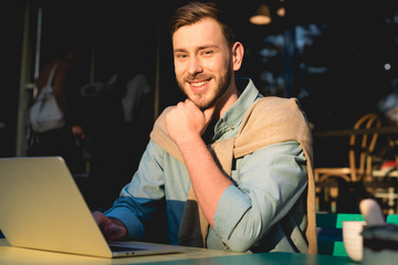 Fototapeta premium cheerful bearded freelancer sitting near laptop and looking at camera