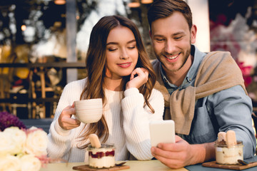 cheerful man looking at smartphone near happy girl holding cup of coffee