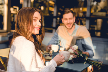 selective focus of happy girl holding cup with coffee while sitting near man in cafe