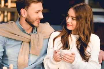 cheerful boyfriend looking at happy girlfriend holding cup of coffee