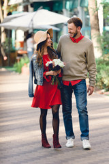 happy man walking with attractive girlfriend in hat holding flowers