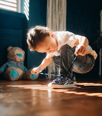 Toddler boy learns to tie the shoelaces on his sneakers. - obrazy, fototapety, plakaty