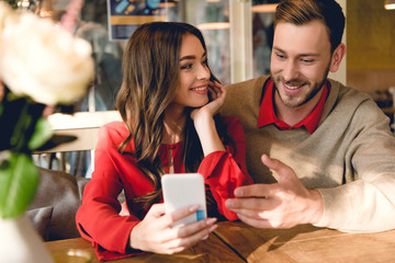 cheerful young woman looking at man gesturing while looking at smartphone
