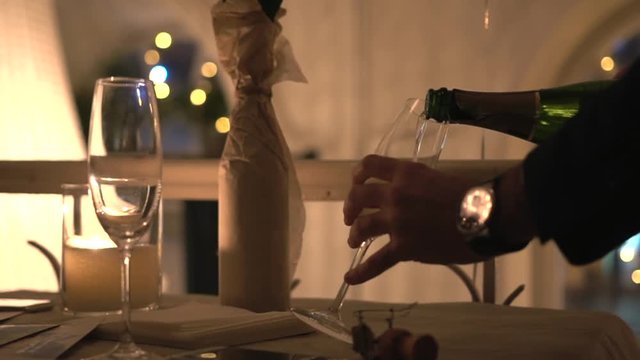 Man in black jacket with arm watch fills a transparent glasses with cold sparkling champagne. White burning candle illuminate bottle covered with craft paper on table in old cafe. Romantic dinner.