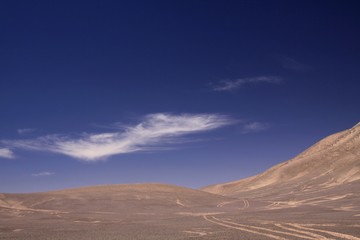 Fototapeta na wymiar Lost in the Landscapes of Atacama desert - Wheel tracks in barren arid sand plain in the nowhere contrasting with deep blue sky and few white cirrus clouds - Chile