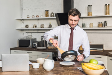 handsome man with frying pan preparing breakfast while standing by kitchen table