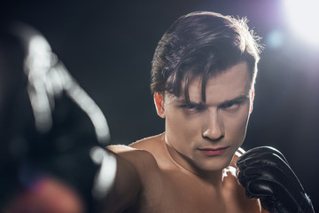 Selective focus of concentrated boxer in boxing gloves training and looking at camera on black