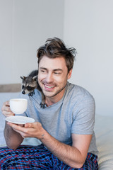 smiling man with funny raccoon on shoulder holding coffee cup while sitting on bedding