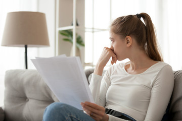 Upset thoughtful woman holding paper document in hands, sitting on sofa