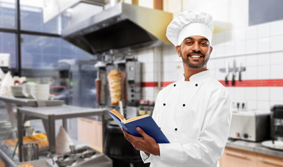 cooking, profession and people concept - happy male indian chef in toque with cookbook over kebab shop kitchen background