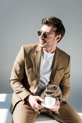 smiling man sitting on sunlight and holding funny raccoon and coffee cup on grey