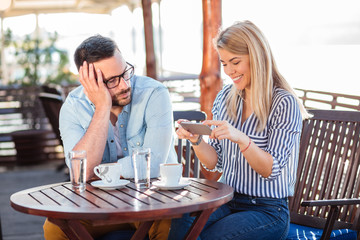 Bored young man waiting for his girlfriend to stop using the phone. Drinking coffee in an outdoor cafe, summertime