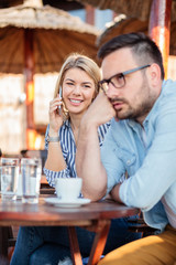Young couple sitting outdoor in a cafe. Woman is talking on the phone and looking at her boyfriend who is bored and annoyed