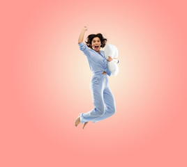 Fototapeta na wymiar fun, people and bedtime concept - happy young woman full of energy in blue pajama holding pillow and jumping over living coral pink background