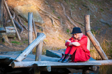 Thoughtfully woman in a coat and hat is sitting by the lake.