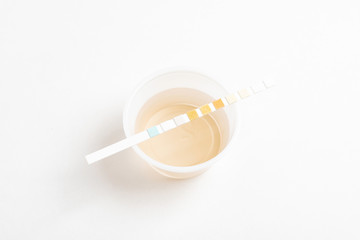 A set of urine sample on a round plastic cup with a single unused urine reagent strip against a...