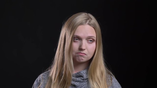 Portrait of young blonde straight-haired model blowing up her cheecks in confusion on black background.