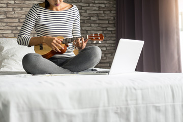 Happy young asian woman playing ukulele sitting on bed in bedroom.