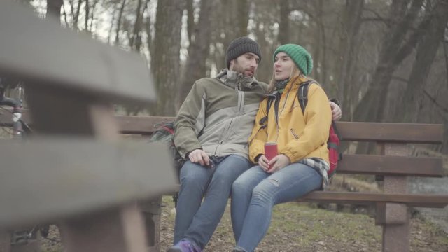 Traveling friends. A young couple of tourists with backpacks sitting on the bench in the spring park at the riverbank. The bearded man and woman in yellow jacket talking. The bicycle standing near