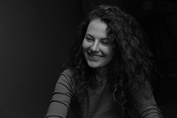 Black and white authentic horizontal portrait of cheerful Caucasian curly haired  female model, smiling, looking aside,having conversation with friend, posing in dark room indoors. Beauty concept.