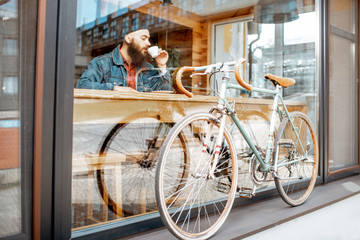 Stylish man enjoying coffee drink while sitting at the cafe near the window with retro bicycle. Wide view through the window