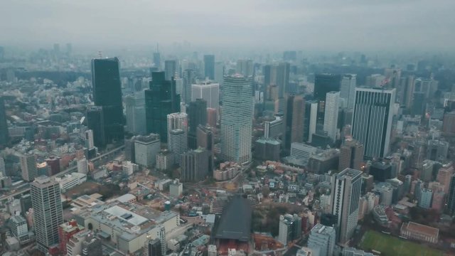Aerial drone panning up away from the Tokyo city skyline in Japan on an overcast day with hazy smog filled skies and grey storm clouds above busy roads and skyscrapers