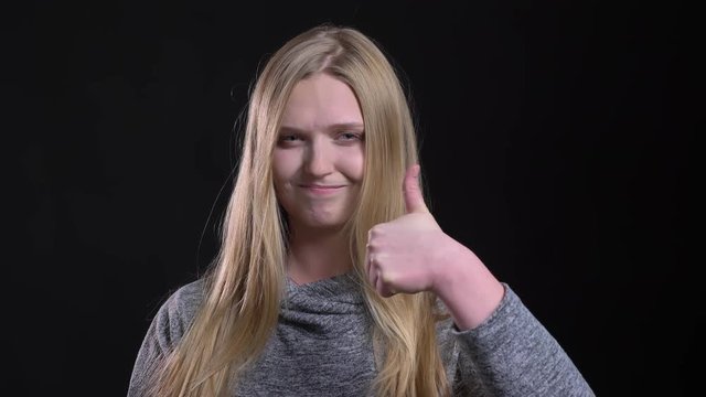 Portrait of blonde straight-haired model gesturing finger-up sign to demonstrate like and respect on black background.