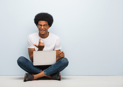 Young black man sitting on the floor with a laptop inviting to come