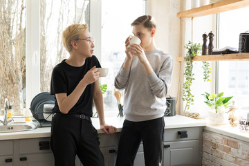 Happy international gay couple at home in casual clothing. Drinking coffee in the kitchen