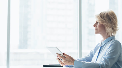 Professional success. Mature female business leader. Side view of blonde woman sitting with tablet against big window.