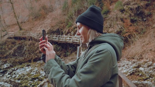 A beautiful blonde girl takes a phone out of the pocket and snaps a photo of a river running through a canyon full of rocks.