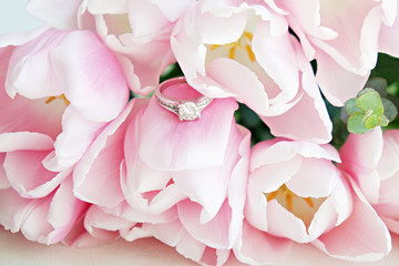 Macro shot of expensive white gold diamond ring and beautiful tender pink tulips. Top view composition with engagement ring and flower petals. Feminine wedding background with copy space for text.