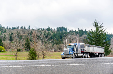Fototapeta na wymiar Blue big rig American classic semi truck transporting cargo in covered bulk semi trailer driving on the flat road