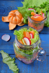 Layered salad in a jar with mandarins, radish, lettuce leaves, carrot and dressing against the blue background