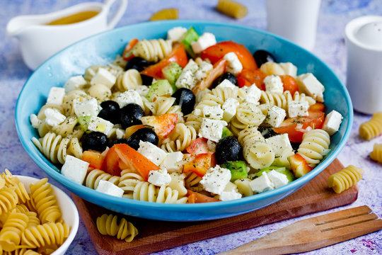 Greek salad with fresh vegetables, feta cheese, pasta and black olives on blue wooden background top view