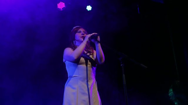 A young red-haired singer sings on stage with professional lighting and puffs of smoke. The average plan