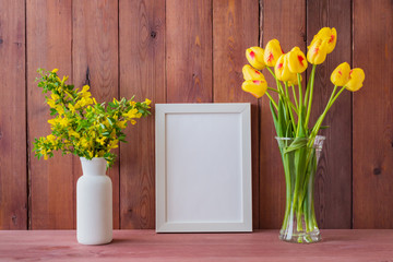 Mockup with a white frame and branches with yellow flowers in a vase on a wooden table